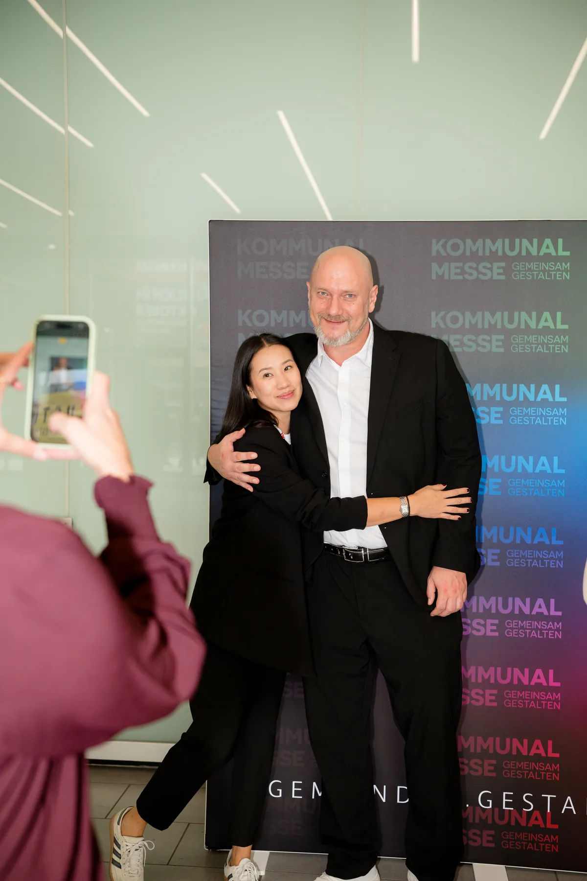 Herzliche Umarmung vor der Fotowand der KOMMUNAL Messe 2026 – vorne wird mit dem Handy mitfotografiert